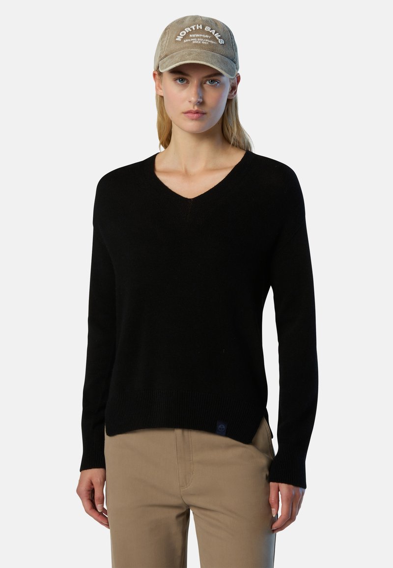 Schwarzer V-Ausschnitt-Pullover aus weichem Material mit geripptem Saum und Bündchen, kombiniert mit einer beigen Cordmütze mit weiß gesticktem Text.