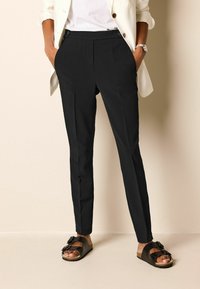 Pantalon noir sur mesure avec une texture lisse, présentant un devant plat, des poches latérales et une coupe slim, associé à des sandales plates foncées.