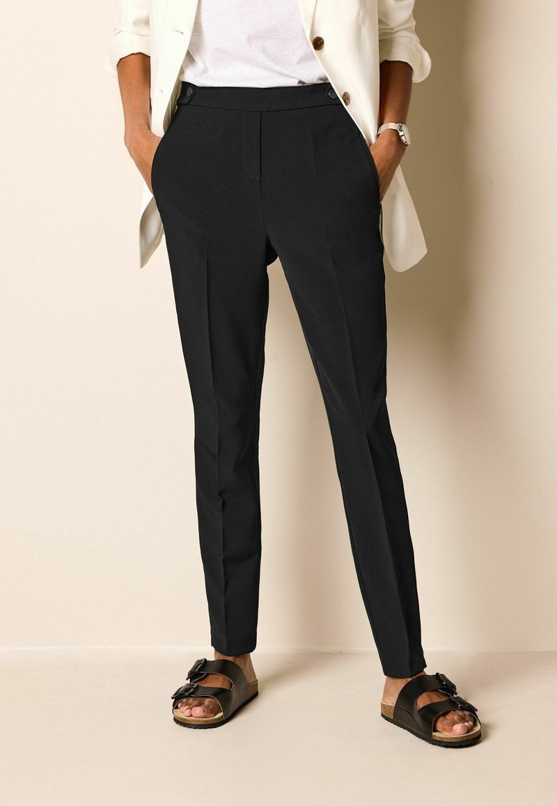 Pantalon noir sur mesure avec une texture lisse, présentant un devant plat, des poches latérales et une coupe slim, associé à des sandales plates foncées.