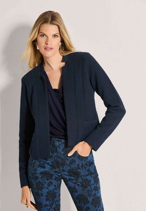 Blonde woman wearing textured dark navy blazer, black top, and blue floral patterned pants, posing with one hand in pocket.