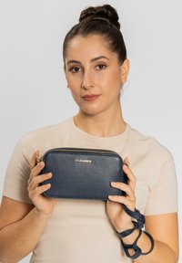 Clutch bag in pelle blu navy con texture a granulato, zip dorata e tracolla staccabile. Presenta un logo impresso sulla parte anteriore.