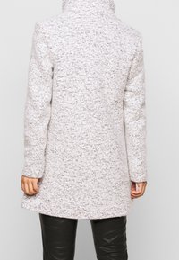 Ljusgrå kappa med ett strukturerat, vävt mönster, hög krage, långa ärmar och knäskärning. Matchad med svarta byxor.