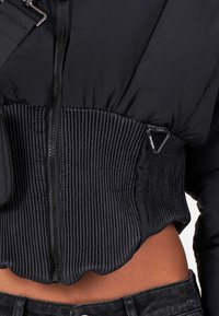 Gros plan sur une veste noire rembourrée avec un détail de taille côtelé, une fermeture éclair à l'avant et un ventre apparent au-dessus d'un jean foncé.