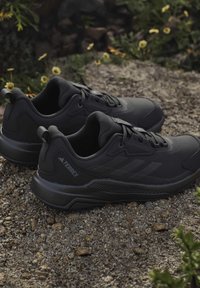 Een paar zwarte Adidas Terrex trail hardloopschoenen op grind met kleine gele bloemen en groen op de achtergrond.