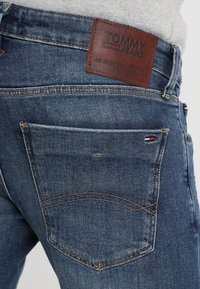 Džínové kalhoty v tmavě modré barvě s koženou nášivkou s nápisem "TOMMY JEANS." Mezi rysy patří zadní kapsy se švy.