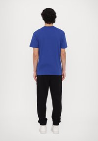 Μπλε βαμβακερό t-shirt με στρογγυλή λαιμόκοψη, κοντά μανίκια και ίσια γραμμή, σε συνδυασμό με μαύρες φόρμες και λευκά αθλητικά παπούτσια. Όψη από πίσω.