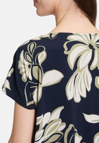 Marinebluse mit großem weißen und olivfarbenen Blumenmuster. Weicher Stoff; kurze Ärmel; runder Ausschnitt; lockere Passform; glatte Textur.