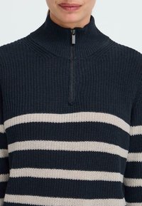 Dunkelblauer Strickpullover mit hohem Kragen und Reißverschluss. Mit horizontalen weißen Streifen und strukturiertem Stoff in lockerer Passform.