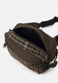 Bolsa de tela marrón con un patrón a cuadros en oliva, azul y negro. Cuenta con un compartimento con cremallera y una correa ajustable.