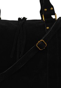 Sac en suède noir avec une texture douce, comportant des accessoires en métal doré, une bandoulière réglable et un détail de frange au niveau de la fermeture.