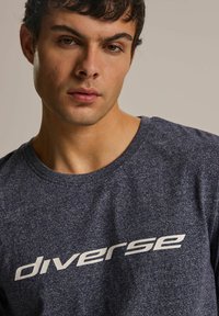 Ciemnoniebieska koszulka z krótkim rękawem, wykonana z teksturowanej tkaniny, z białym nadrukiem logo "diverse" na piersi oraz okrągłym dekoltem.