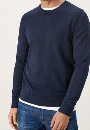 Mężczyzna w granatowym swetrze z długim rękawem i okrągłym dekoltem, noszący biały shirt oraz niebieskie dżinsy, stojący na tle jasnego, jednolitego tła.