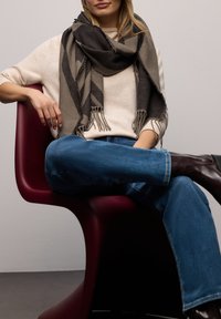 Écharpe à carreaux gris et beige drapée sur un pull crème, associée à un jean bleu et des bottes marron foncé, assise sur une chaise bordeaux.