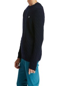 Pull bleu marine en maille torsadée avec poignets et ourlet côtelés, arborant un petit logo sur la poitrine. Conçu avec un col rond et des manches longues.