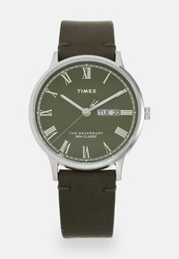 Montre Timex avec un cadran analogique vert, des chiffres romains, un boîtier argenté et un bracelet en cuir vert foncé. Affiche la date et le jour de la semaine.