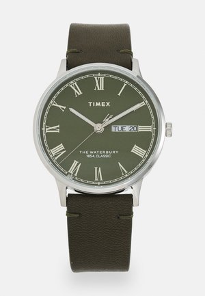 Montre Timex avec un cadran analogique vert, des chiffres romains, un boîtier argenté et un bracelet en cuir vert foncé. Affiche la date et le jour de la semaine.