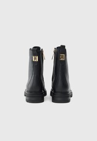 Botas de tobillo de cuero negro con suelas gruesas, etiquetas con logotipos dorados en las lengüetas traseras y cremalleras doradas en los lados interiores, vistas desde atrás.