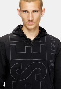 Veste noire à capuche avec une finition texturée, comportant un grand motif imprimé en gris foncé. Comprend des cordons de serrage et une fermeture éclair.