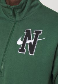 Tmavě zelená mikina na zip s vysokým límcem. Obsahuje velké černé logo "N" a bílé logo Nike, vyšívané pro texturu.