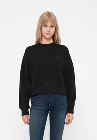 Svart cropped sweatshirt i myk stoff, med rund hals, senkede skuldre og liten rød logo på venstre bryst. Brukt med blå jeans.