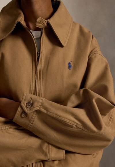 Persona che indossa una giacca color sabbia con cerniera e un logo ricamato di un giocatore di polo blu sul petto, con le braccia incrociate.