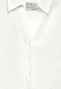 Camisa blanca de botones con tejido texturizado y cuello, que presenta un escote en V profundo y un sutil logotipo en la etiqueta interior.