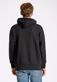 Sudadera de algodón negra con una gran capucha, hombros caídos y puños de canalé; mostrada desde la espalda sobre un fondo claro.