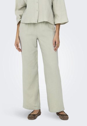 Femme portant un pantalon ample texturé vert clair et une chemise boutonnée assortie, associée à des ballerines marron.