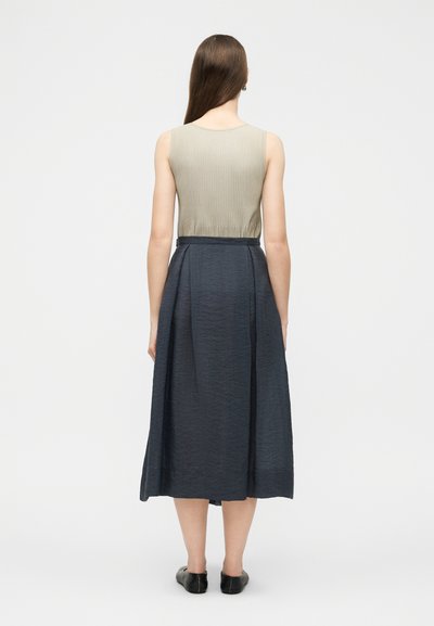 Γυναίκα με μακριά καστανά μαλλιά στέκεται πλάτη, φοράει μπεζ αμάνικο τοπ, μπλε σκούρα μεσάτη φούστα και μαύρα μοκασίνια.