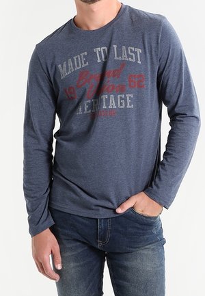 Mężczyzna w niebieskiej koszuli z długim rękawem z napisem „Made to Last 1962 Brand Vision Heritage”, w połączeniu z niebieskimi dżinsami, ręka w kieszeni.