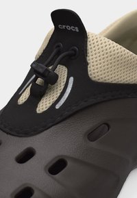 Κοντινό πλάνο ενός μαύρου και μπεζ παπουτσιού Crocs με υφή στην επάνω επιφάνεια, κορδόνια με κλείδωμα και οπές αερισμού στη σόλα.