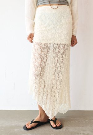 Maxi-Rock aus Spitze in Creme mit transparentem Design, verziert mit blumigen Mustern, einem fließenden Saum und einem eng anliegenden Taillenbund. Kombiniert mit schwarzen Flip-Flops.