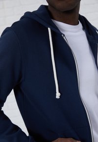 Marinblå zip-up hoodie med vita dragsnören. Har en framficka och en mjuk textur, bärs över en vit t-shirt.