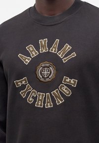Μαύρο φούτερ με μπεζ κεντημένο κείμενο "Armani Exchange" και κυκλικό έμβλημα στη μέση με την επιγραφή "Milano A/X 91 Est. 1991".