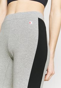Leggings grigi con pannello laterale nero, realizzati in tessuto morbido, presentano un piccolo logo sulla cintura. Texture liscia con design aderente.