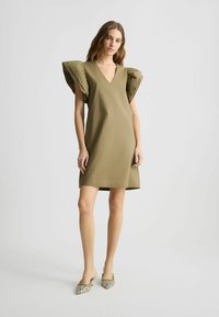 Khaki haljina s dekolteom u obliku slova V i oversized volanima. Glatka tkanina, dužina iznad koljena. Uparena s cipelama na špic s uzorkom.