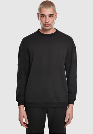 Must ülipikk dressipluus ümmarguse kaelusega, pikkade varrukatega ja külgedele paigutatud tõmblukkudega taskutega. Valmistatud pehmest materjalist, millel on soonikkoes mansetid ja allserv.