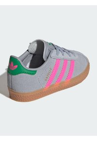 Pilko zomšos sportiniai bateliai su rožinėmis trimis juostelėmis, žaliu kulno akcentu ir dantytu guminiu padu. Pagaminti su paminkštintu apykakle ir auksiniu prekės ženklu.