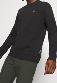Hombre vistiendo una sudadera negra de manga larga con un pequeño emblema de la bandera noruega y pantalones verde olivo, de pie contra un fondo sencillo.