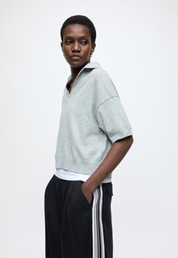 Svetlo sivi pulover s kratkimi rokavi in ovratnikom, izdelan iz mehke tkanine. Kombiniran s črnimi širokimi hlačami z belimi stranskimi črtami.