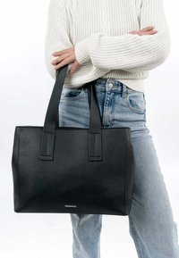 Zwarte leren tote bag met een gestructureerd oppervlak, twee stevige handvaten en een eenvoudig ontwerp, vastgehouden door een persoon in een witte sweater en blauwe jeans.