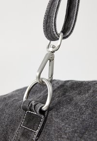 Alça de bolsa de tecido cinza com superfície texturizada, apresentando um clipe de metal prateado e um anel circular para fixação. Detalhes de costura são visíveis.