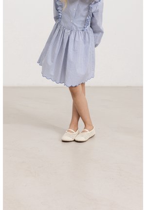 Robe rayée bleu et blanche avec des bords volants, longueur genou. Chaussures blanches à enfiler en tressé avec deux brides sur le dessus. Fond neutre.