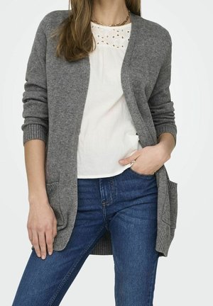 Frau, die ein weißes Lochstick-Top, blaue Jeans und eine graue Strickjacke mit Taschen trägt, eine Hand in der Tasche, dargestellt vom Hals bis zu den Oberschenkeln.