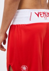 Ανδρικός κορμός με κόκκινα σορτς Venum, λευκή ελαστική μέση και λογότυπο κοντά στο αριστερό πόδι, στέκεται με το αριστερό χέρι χαλαρό στο πλάι.