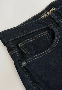 Jeans de mezclilla oscura con un bolsillo frontal, costuras visibles y un acento de botón. La tela tiene una textura suave con un ligero brillo.