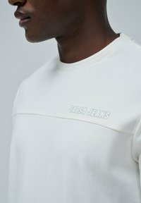 Sudadera blanca hecha de una tela suave, con cuello de canalé, costura horizontal y logo "SALSA JEANS" en gris claro a lo largo del pecho.