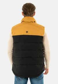 Pufferväst med en gul överdel och svart underdel, med horisontell söm, hög krage och en logotyp på baksidan.