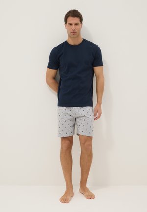 Man standing barefoot against wall wearing navy blue short-sleeve shirt and light gray shorts with small anchor and sailors hat patterns.