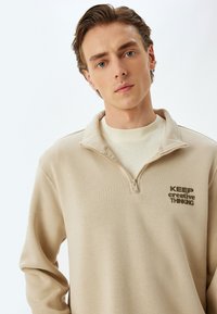 Beiger Pullover mit einem Reißverschlusskragen, ausgestattet mit einem strukturierten Stoff und bedrucktem Text "HALTE kreatives DENKEN" in Dunkelbraun auf der Vorderseite.